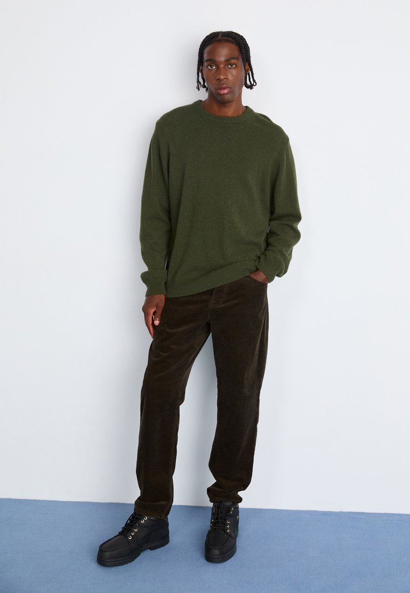 Grüner Strickpullover mit gerippten Bündchen, kombiniert mit dunkelbraunen Cordhosen. Schwarze Schnürstiefel mit goldenen Ösen vervollständigen das Outfit.