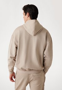 Felpa beige con una vestibilità morbida, dotata di cappuccio e polsini a costine. Texture morbida con cuciture evidenti, abbinata a pantaloni coordinati.