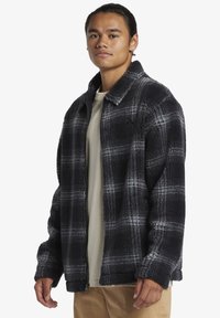 Black and gray plaid fleece jacket with a pointed collar, zip closure, and side pockets. Soft texture, relaxed fit, and long sleeves.