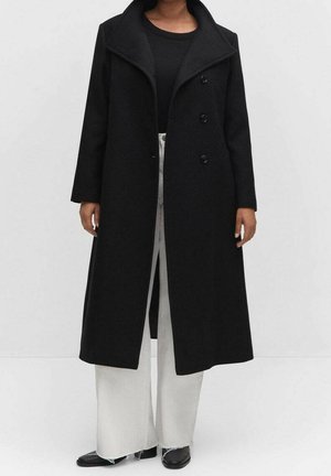 Mujer con un abrigo largo negro de doble botonadura, top negro, pantalones blancos de pierna ancha y zapatos negros sobre un fondo blanco liso.