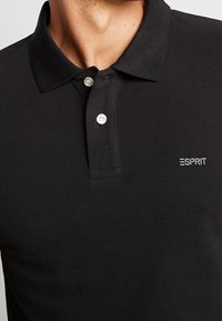 Polo noir en tissu texturé, doté d'un col classique, de deux boutons blancs et d'un petit logo Esprit blanc sur la poitrine.