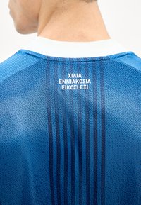 Homme portant un maillot de sport bleu avec un col blanc, avec un texte grec imprimé en blanc sur le haut du dos sur un fond blanc.