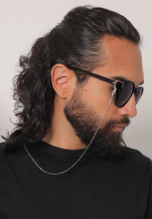 Mann mit langen Haaren und Bart, der schwarze Sonnenbrillen trägt, die an einer dünnen silbernen Kette hängen, gekleidet in ein schwarzes Hemd, und nach rechts schaut.