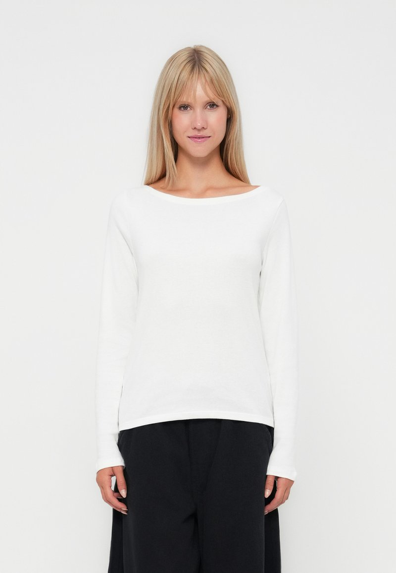 Camiseta blanca de manga larga con un amplio escote barco, hecha de una tela suave. Presenta un corte holgado y se sitúa a la altura de las caderas. Combinada con pantalones oscuros.