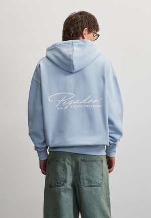 Homme portant un sweat à capuche bleu clair avec le texte « Pegador ALL RIGHTS RESERVED » dans le dos et un pantalon vert ample, tourné de dos sur un fond uni.