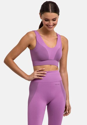 Vrouw in een bijpassende lavendelkleurige sportbeha en hoge taille legging, glimlachend met hand op de heup tegen een effen achtergrond.