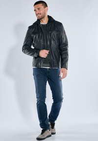 Black leather jacket with a hood, front zippers, two chest pockets, textured finish, paired with blue jeans and beige high-top sneakers.