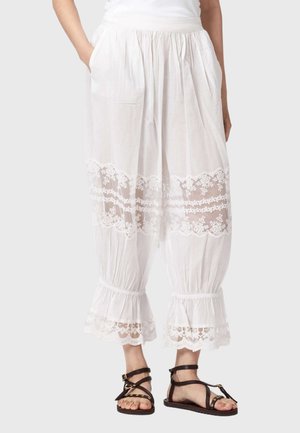 Kobieta nosząca białe, luźne spodnie do połowy łydki z koronkowymi panelami i falbanami na nogawkach, zestawione z czarnymi, płaskimi sandałami na paski.