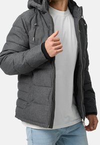 Man i grå vadderad jacka med delvis uppladdad fram över vit t-shirt och ljusblå jeans, justerar jackans krage.