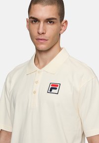 Camisa polo crema de algodón, con una tapeta de tres botones y un logo bordado en rojo, blanco y azul en el lado izquierdo del pecho.