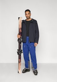 Roupa de esqui composta por um casaco impermeável azul-marinho, camisa preta e calças de esqui azuis com detalhes cinzentos. A segurar esquis castanhos com fixações pretas.