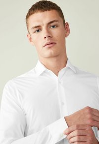 Cămașă albă cu mâneci lungi, cu un guler ascuțit și închidere cu nasturi. Textură netedă, design ajustat, evidențiat cu o manșetă butonată și un stil minimalist.