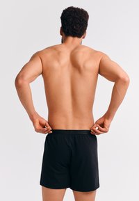 Fit man met krullend haar past de tailleband van zwarte boxershorts aan, gezien van achteren op een effen lichte achtergrond.