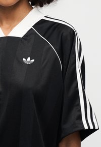 Μαύρο αθλητικό πουκάμισο με λευκές λεπτομέρειες, με V-λαιμόκοψη, κοντά μανίκια και τρεις λευκές ρίγες στις πλευρές. Το λογότυπο της Adidas εμφανίζεται ξεκάθαρα.