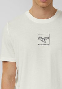 Koszulka z krótkim rękawem, wykonana z jasnobeżowej bawełny, z kwadratowym nadrukiem przedstawiającym czarne linie artystyczne oraz napisem „Elevated Casual” pod spodem.