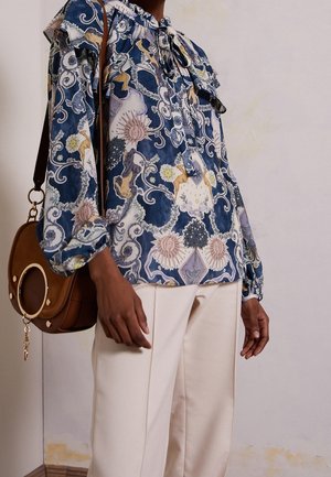 Osoba oblečená v modré blůze s motivy slunce a květin, krémových kalhotách a nesoucí hnědou koženou kabelku přes rameno se zlatým kroužkem.