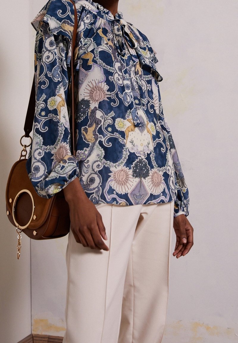 Personne portant un chemisier bleu à motifs de soleil et fleurs, un pantalon crème, et un sac à bandoulière en cuir marron avec un anneau doré.