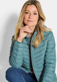 Teal pufferjack met horizontale gesteven patroon, volledige ritssluiting en opstaande kraag. Gecombineerd met een beige top en blauwe denim jeans.