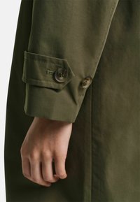 Tela de algodón verde oliva con un puño que cuenta con dos botones marrones y una solapa decorativa. Se pueden apreciar la textura suave y la forma entallada.