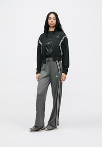 Sieviete stāv melnā un pelēkā Jordan treniņtērpā ar baltām svītrām un pelēkām sporta kurpēm uz vienkārša gaiša fona.