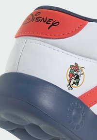 Sneaker bianca con accenti rossi e blu navy. Presenta un logo Disney e un grafico di personaggi dei cartoni animati sul tallone. Texture liscia, suola rotonda.