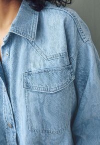 Camicia in denim blu chiaro con colletto a punta, bottoni a pressione argentati e taschino sul petto. Il tessuto ha una texture leggermente invecchiata.