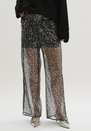 Pantalon noir transparent orné de sequins argentés, design à jambes larges, tissu texturé, porté avec un haut noir et des ballerines argentées.