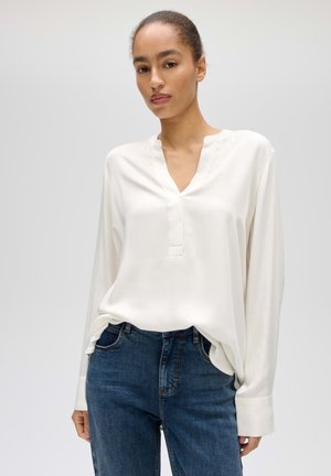 Donna con i capelli raccolti che indossa una camicetta bianca larga a maniche lunghe e jeans blu, in piedi davanti a uno sfondo chiaro e uniforme.
