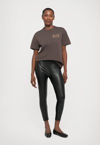 Brun bomull t-skjorte med korte ermer og gulllogo, kombinert med svarte glinsende faux skinn leggings og svarte flate sko.