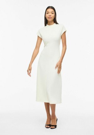 Kobieta stoi w białej sukience midi z krótkimi rękawami i stójką, ma na sobie czarne buty na obcasie z odkrytymi palcami na jednolitym białym tle.