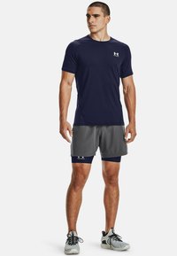 Marineblaue Sport-T-Shirt, kurze Ärmel, leichtes Material; kombiniert mit grauen Shorts und navy blauen Kompressionsshorts darunter. Graue Laufschuhe.