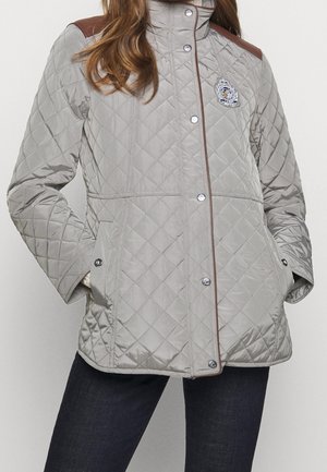 Vrouw draagt een lichtgrijze doorgestikte jas met drukknopen, bruine schouderaccenten, een geborduurd wapen en haar handen in de zakken.