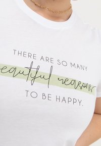 Vit bomullströja med svart text i handskriven stil och en ljusgrön accentlinje. Texten lyder: "DET FINNS SÅ MÅNGA vackra anledningar TILL ATT VARA GLAD."