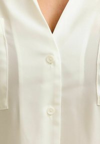 Närbild på vit blus med två synliga knappar och en V-ringning, som visar en del av nyckelbenet och sömmen fram.