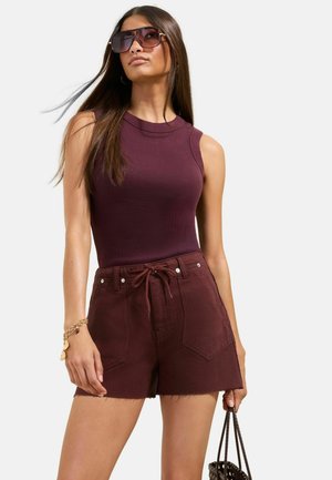 Frau trägt ärmelloses, dunkelburgunderfarbenes Oberteil, passende hoch taillierte Shorts, Sonnenbrille, Armband, hält eine geflochtene Tasche an ihrer Seite.