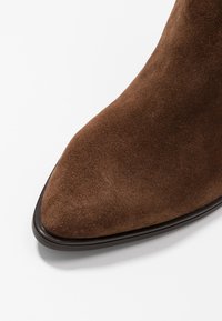 Hnědé semišové kotníkové boty s špičatou špičkou, elegantním designem a hladkou tmavou podešví. Textura je jemná, ukazující minimalistickou estetiku.