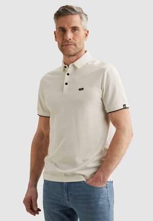 Mann mit grauen Haaren, der ein cremefarbenes Poloshirt mit schwarzen Knöpfen und blaue Jeans trägt, steht mit einer Hand in der Tasche vor einfarbigem Hintergrund.
