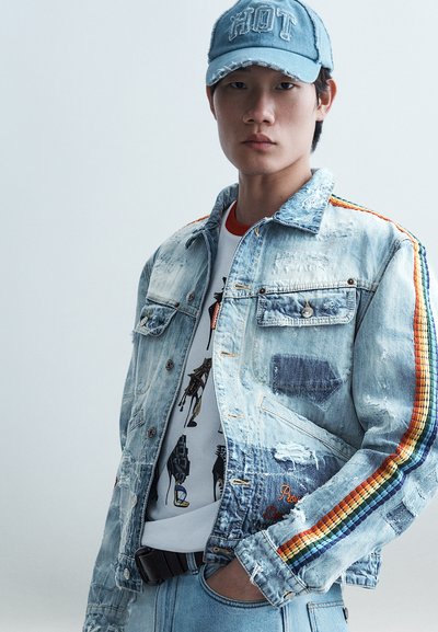 Jeune homme portant une veste en jean usée avec des bandes arc-en-ciel sur les manches, un t-shirt graphique, un jean clair et une casquette bleue avec le texte "HOT".