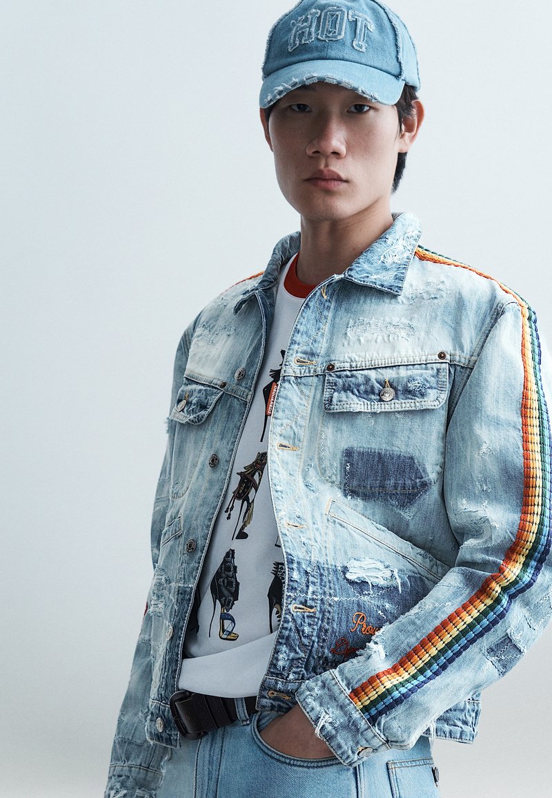 Νέος άνδρας που φοράει ένα φθαρμένο τζιν μπουφάν με ρίγες ουράνιου τόξου στα μανίκια, γραφικό μπλουζάκι, ανοιχτόχρωμο τζιν και μπλε καπέλο με το κείμενο "HOT".