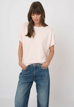 Kobieta z długimi brązowymi włosami, ubrana w luźny blady różowy top z krótkim rękawem i wysokim stanem szerokie niebieskie dżinsy z rękami w kieszeniach.