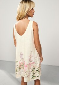 Robe sans manches de couleur crème avec un dos en V, ornée d'un imprimé floral rose et vert à l'ourlet, fabriquée dans un tissu léger.