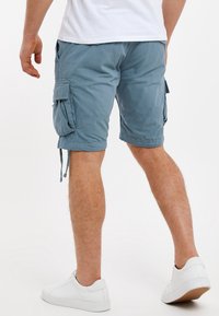 Muž v světle modrých cargo šortkách a bílých teniskách, stojící na bílém pozadí, z pohledu zezadu.