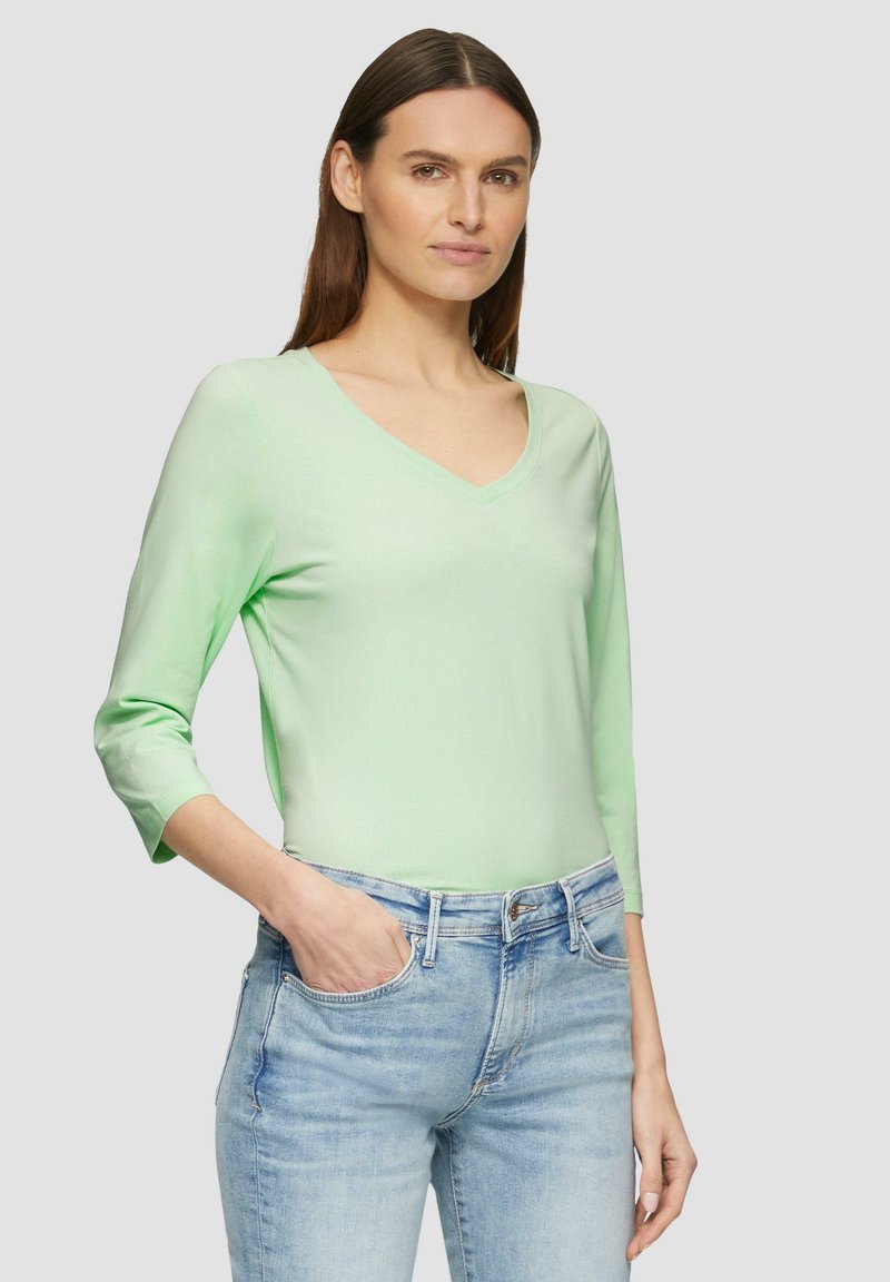 Lichtgroene lange mouwen top met een V-hals, gemaakt van zachte stof. Gemaakt met lichtblauwe denim jeans, staande tegen een neutrale achtergrond.