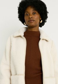 Chaqueta de forro polar color crema con cierre frontal y cuello, que cuenta con bolsillos laterales. Lleva puesta una chaqueta de cuello alto de canalé marrón.