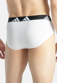 Weiße Baumwollslips mit einem schwarzen Bund, der mit weißen Adidas-Streifen verziert ist. Das Design hebt eine enge Passform und eine glatte Textur hervor.
