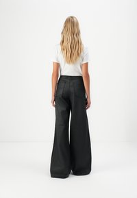 Femme blonde portant un haut blanc à manches courtes et un pantalon noir large taille haute, se tenant face à un fond blanc uni.