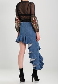 Gonna in denim blu asimmetrica con orlo arricciato; vita alta e bordi sfrangiati; abbinata a un top nero trasparente e stivali alla caviglia.