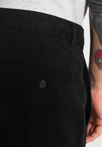 I pantaloni di cotone neri presentano una superficie liscia, una tasca posteriore con un bottone nero e dettagli di cucitura lungo la vita.