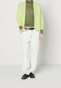 Cardigan verde chiaro oversize con trama a costine, indossato sopra un dolcevita aderente verde scuro, abbinato a jeans bianchi larghi e mocassini neri.