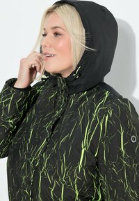 Kvinde med blondt hår iført en sort hættejakke med neongrønne abstrakte linjer, der retter på sit hår nær hætten.